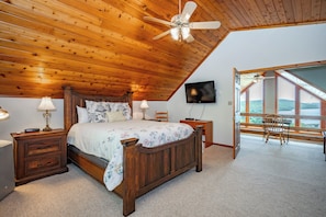 Une chambre avec un plafond en bois, un lit recouvert d’une literie à motifs floraux, une télévision et une vue dégagée grâce à de grandes fenêtres.