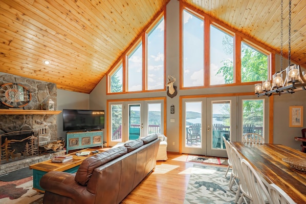 A spacious living room with a high ceiling, large windows, a stone fireplace, a leather sofa, and a wooden dining table.