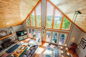 Un salon chaleureux avec une cheminée en pierre, un grand téléviseur et une vue sur l’extérieur grâce à de grandes fenêtres.