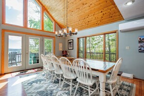 A dining area with a wooden table, white chairs, and large windows with a view of trees.