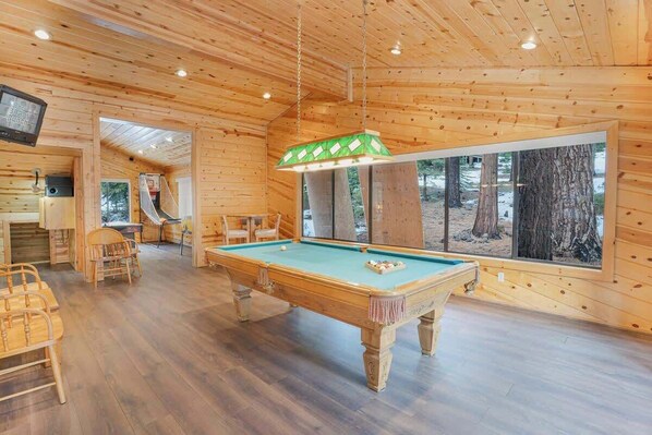 A wooden interior with a pool table, chairs, a television, and a large window.