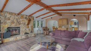 Un salon avec un mur en pierre, des poutres en bois, une cheminée, un canapé violet et une table basse en verre.