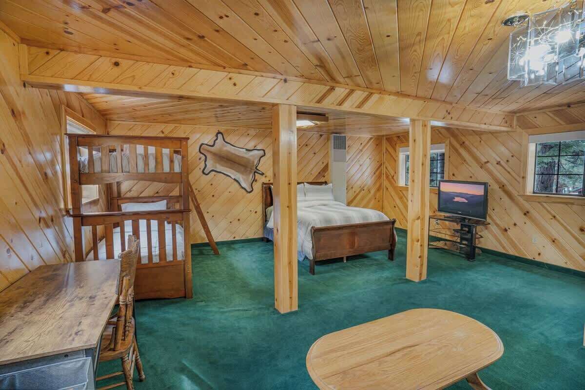 Interior de una cabaña con una cama, un escritorio, una silla, un televisor y un ventanal grande.
