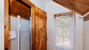Ein Badezimmer aus Holz mit einer Duschkabinentür aus Glas, einem weißen Handtuch und einem Fenster mit durchsichtigem Vorhang.
