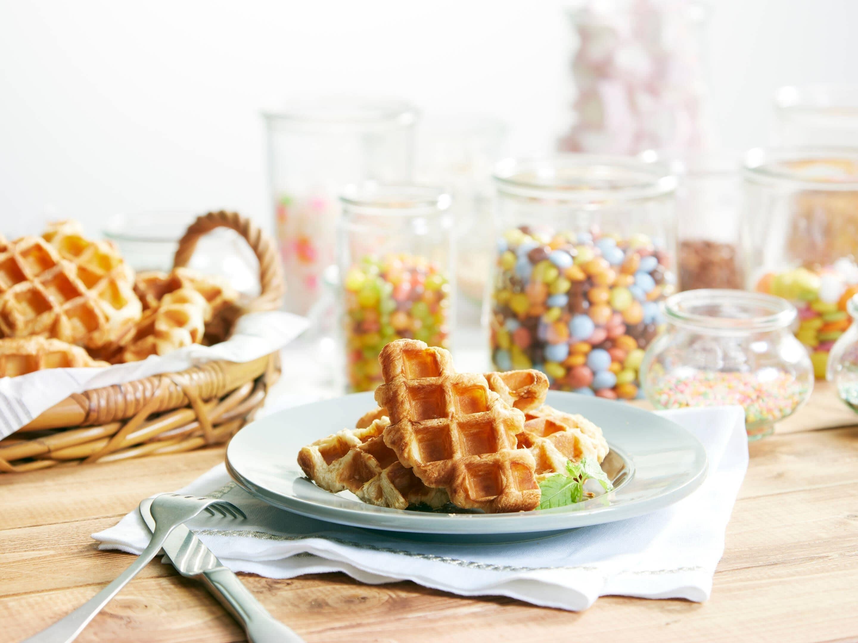 Waffles on a plate with a fork, a basket, and jars of candies in the background.