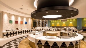 Um hall de hotel moderno com um balcão central redondo, diversas áreas de estar e uma parede decorativa com padrões geométricos.