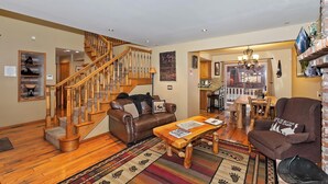 A spacious living room with a wooden staircase, a dining area with a chandelier, and a kitchen visible through an open door. - HOT TUB! PET FRIENDLY! Two Game Rooms! Propane BBQ! Log Furnishings! WiFi! (Big Bear City)