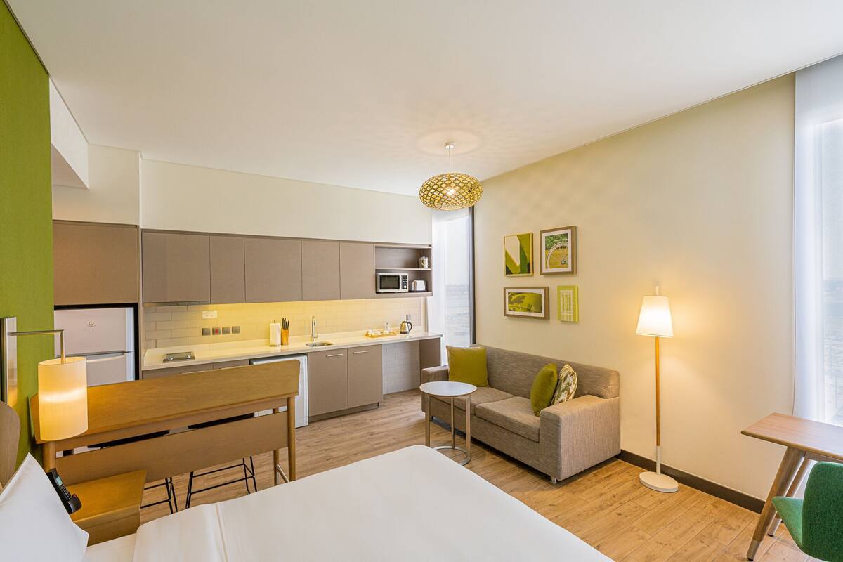 Ein kompaktes Hotelzimmer mit Küchenzeile, einer Couch, einem Esstisch und einem Nachttisch.