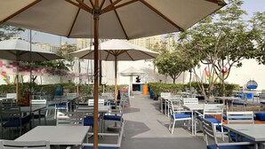 Un café en plein air avec des tables blanches et des chaises bleues dotées de coussins, à l’ombre de grands parasols, le tout sur fond de bâtiments et d’arbres.