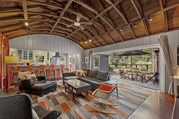 A spacious living area with a large sofa, a coffee table, and a rocking chair. There is a kitchen area with bar stools and a dining area with a table and chairs. The space has a high ceiling with wooden beams and a ceiling fan.