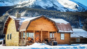 A wooden cabin with a porch and a chimney, surrounded by snow-covered mountains. - Log Cabin on Slate River | 4 Bedroom | Mt CB View | River Access (Crested Butte)