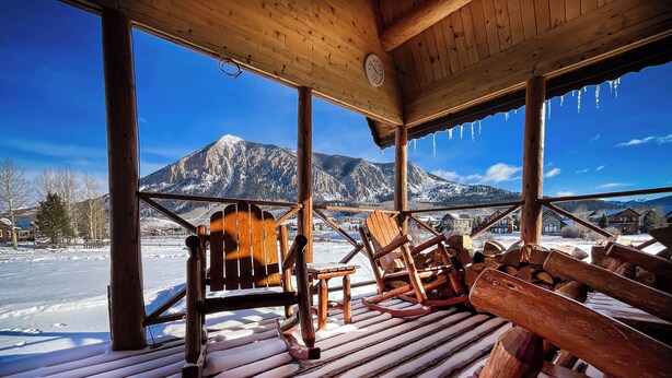 A wooden deck with Adirondack chairs and a snow-covered ground, overlooking a mountainous landscape. - Log Cabin on Slate River | 4 Bedroom | Mt CB View | River Access (Crested Butte)