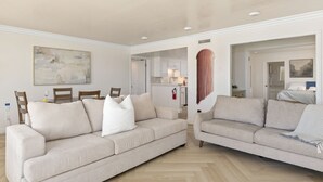 Una sala de estar con dos sofás color beige, una mesa de comedor con sillas, una zona de cocina y un pasillo que conduce a un dormitorio.