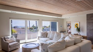 A spacious living room with a large white sofa, a smaller armchair, and a round coffee table. The room features a wooden ceiling and large sliding glass doors leading to a balcony with a view of the sea. - Luxury 2 Bed/2 Bath Penthouse in Shell Beach, Overlooking the Coast with Wrap-Around Balconies (Shell Beach)