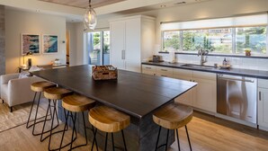 A modern kitchen with a large island, bar stools, stainless steel appliances, and a view through a large window. - Luxury 2 Bed/2 Bath Penthouse in Shell Beach, Overlooking the Coast with Wrap-Around Balconies (Shell Beach)