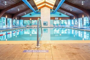 Piscina interna com um aviso de 'NÃO MERGULHAR', vigas de madeira e grandes janelas.