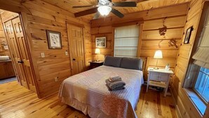 Un dormitori de cabana de fusta amb un llit, dues taules de nit, un ventilador de sostre i una finestra.
