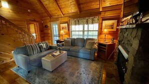 Interior acollidor d'una cabana de fusta amb sostre de fusta, una llar de pedra, una catifa blava amb dibuixos i dos sofàs.