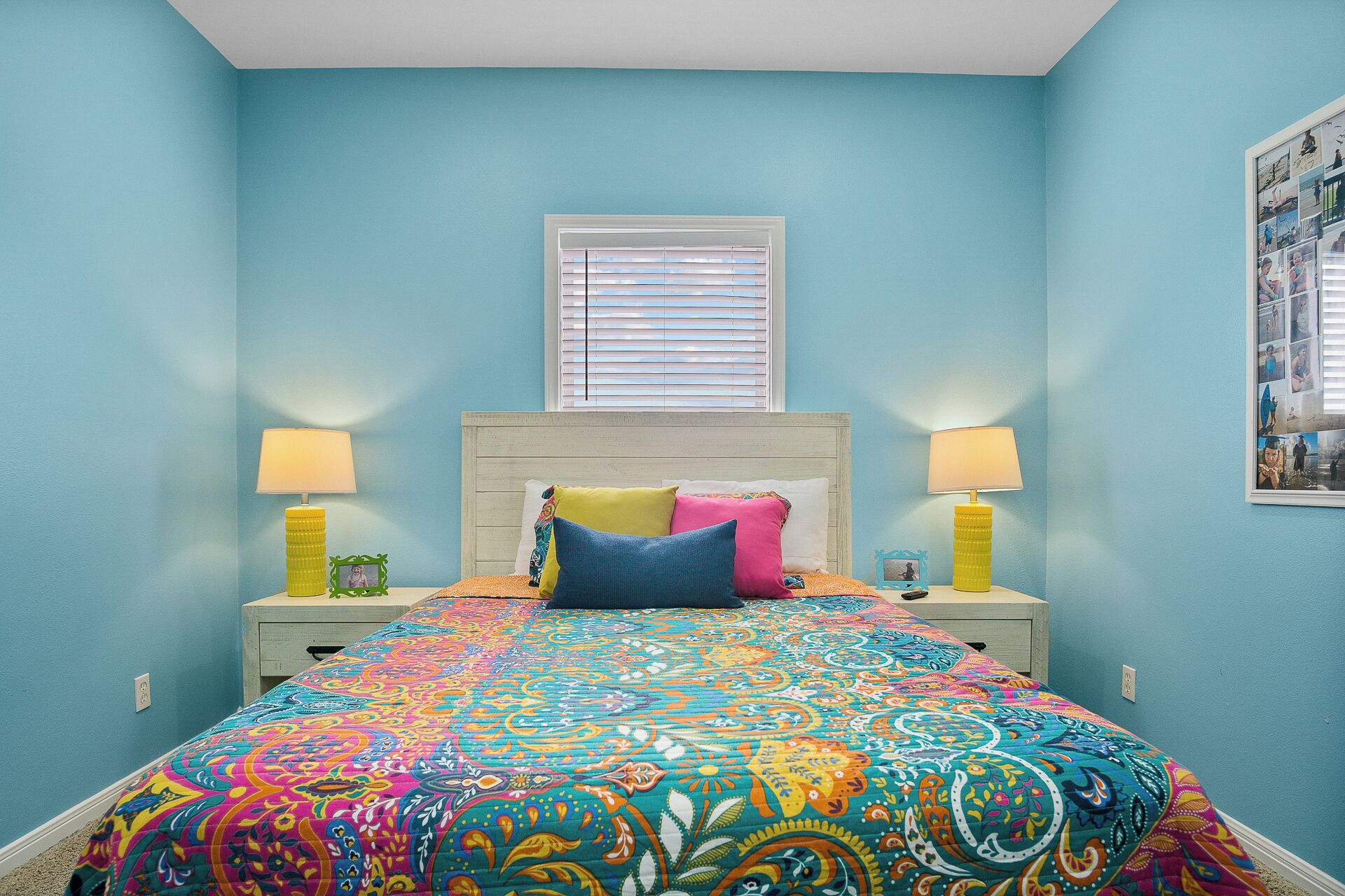 A bedroom with a bed, two bedside lamps, a window with blinds, and a framed collage on the wall.
