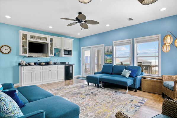 Uma sala moderna com sofá azul, área de jantar com armários brancos, cozinha com micro-ondas embutido e grandes janelas com vista.