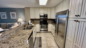 Een moderne keuken met witte kasten, roestvrijstalen apparatuur, een granieten aanrechtblad en uitzicht op de woonkamer.