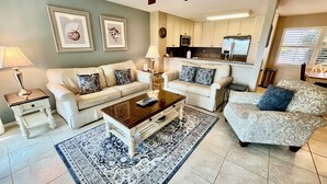 Een woonkamer met een bank, salontafel en een vloerkleed met patroon. Achtergrond is een keuken met kasten en apparatuur.