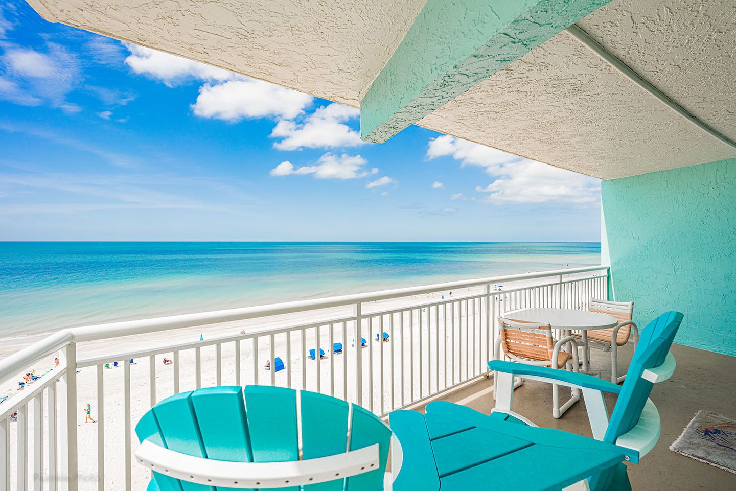 Un balcon avec des chaises turquoise et une table, donnant sur une plage aux eaux d’un bleu limpide et bordée d’une rambarde blanche.