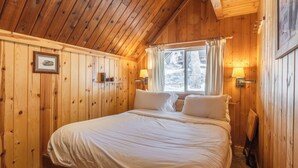 A wooden cabin bedroom with a bed, a window with curtains, a wall-mounted lamp, and a shelf with a picture and an apple. - Chalet #2, Charming Ski In/ Ski Out Steps to the M (Mammoth Lakes)