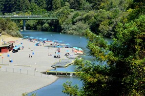 Ein Flussstrand mit Kanus, Sonnenschirmen und einer Brücke im Hintergrund.