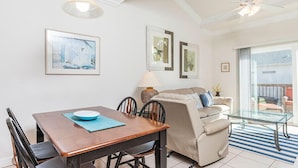 Un comedor con una mesa de madera, sillas y un mantel azul. En la zona de estar, hay un sofá color beige y una mesa de centro de cristal. La habitación tiene obras de arte enmarcadas en las paredes y un ventilador de techo.