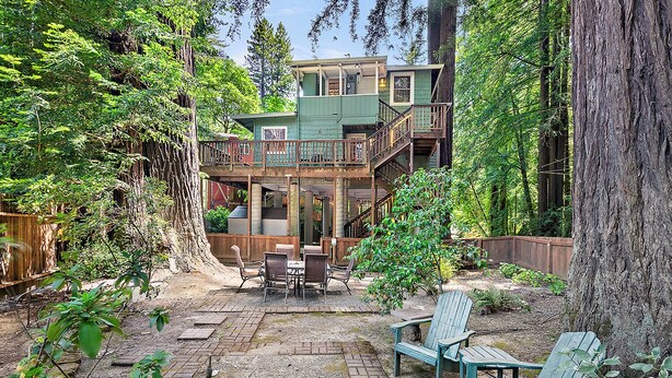 A two-story wooden cabin with a deck, surrounded by dense forest. - Monte Rio Treehouse ~ Dog Friendly, Redwoods, Walk to Restaurants and River (Monte Rio)