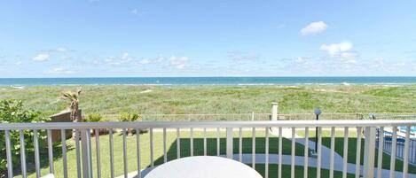A balcony with a white table and chairs, overlooking a beach with a clear sky.