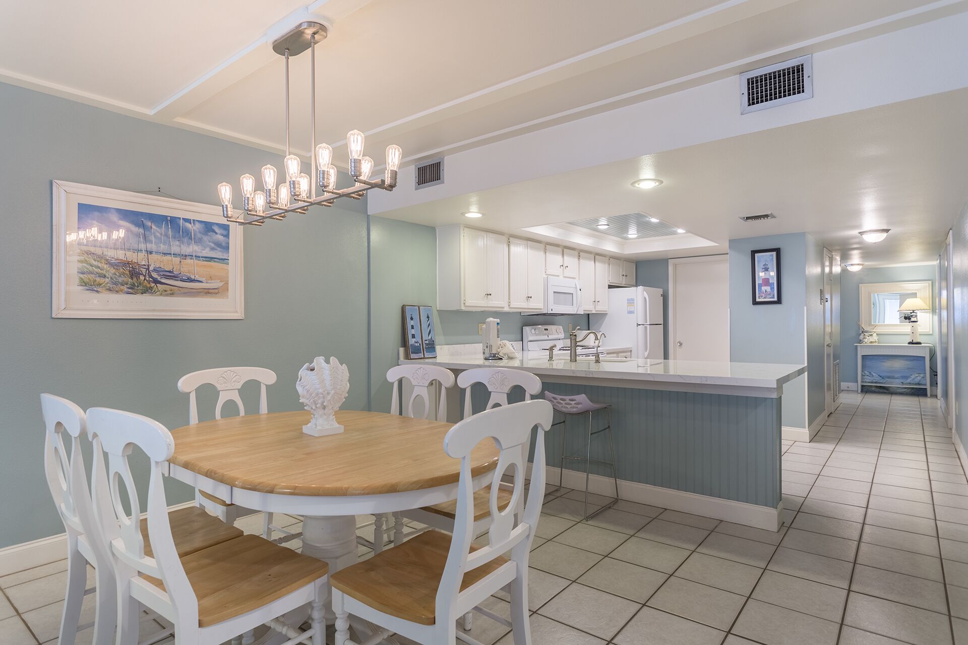 A dining area with a round wooden table and white chairs, a kitchen with white cabinets, and a hallway with a blue accent wall.