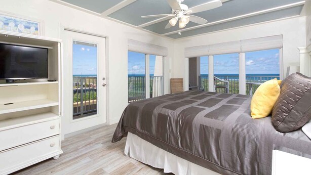 A bedroom with a bed, a TV, a ceiling fan, and large windows with a view of the ocean.