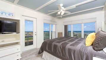 Un dormitorio con cama, televisión, ventilador de techo y ventanas amplias con vista al océano.
