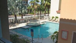 Piscina com pequena área de deck, cercada por palmeiras e um edifício com telhado ladrilhado.