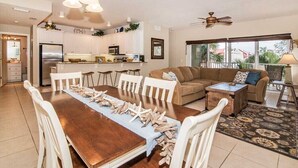 Uma sala de estar espaçosa com uma grande mesa de jantar de madeira, cadeiras brancas, um sofá marrom e um ventilador de teto.