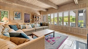 A spacious living room with a large sectional sofa, a coffee table, and a rug. There are large windows providing a view of the outdoors. - Classic Second Row Beach House, Short Walk to the Beach (Georgetown)