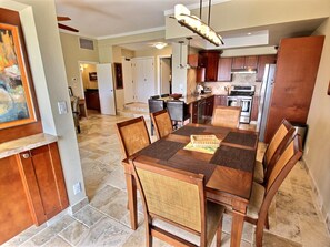A spacious dining area with a large wooden table and chairs, a kitchen in the background, and a hallway leading to another room.