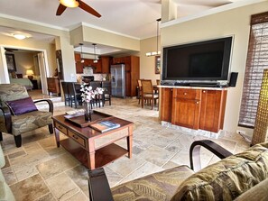 A spacious living room with a large flat-screen TV, a dining area with wooden chairs, and a kitchen in the background.