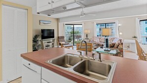 A modern kitchen with a double sink, a flat-screen TV, and a dining area with a glass table and chairs. - Water View 207 - INTERCOASTAL VIEW Condo! (Indian Shores)