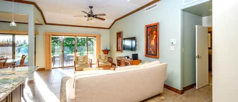 A spacious living room with a large sofa, a dining area with a table and chairs, a kitchen area with a countertop, and a ceiling fan.