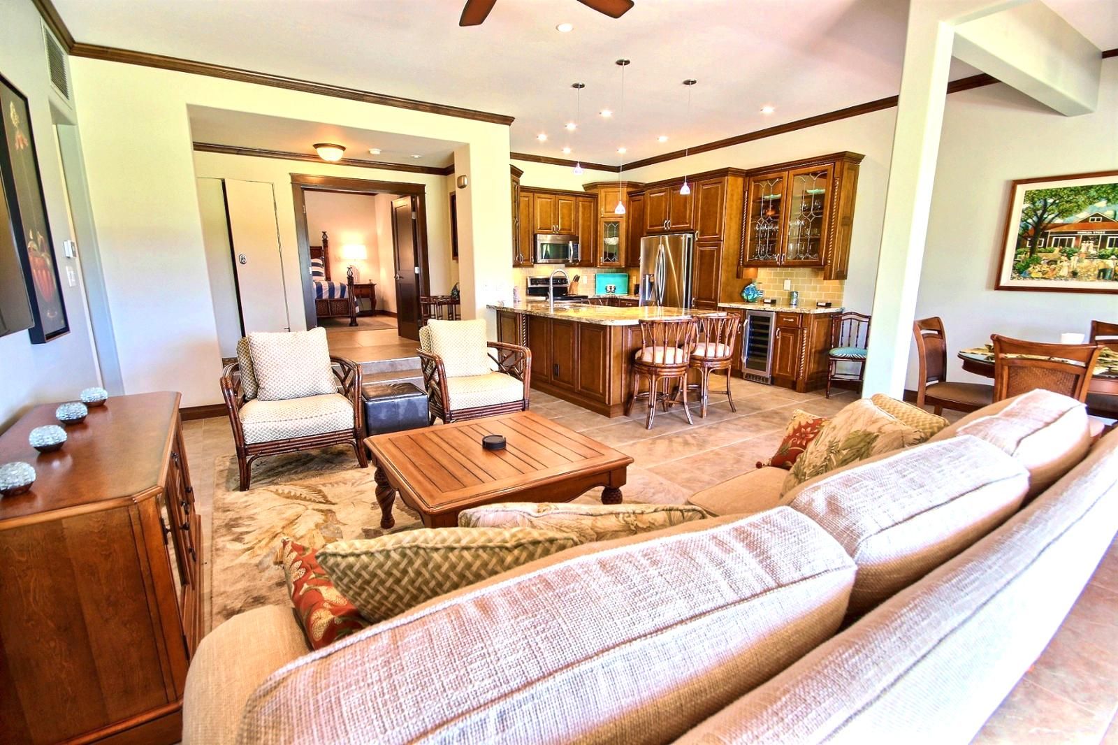 A spacious living room with a large sofa, a coffee table, and a dining area with chairs. There is a kitchen area with wooden cabinets and a refrigerator.
