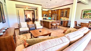 A spacious living room with a large sofa, a coffee table, and a dining area with chairs. There is a kitchen area with wooden cabinets and a refrigerator.