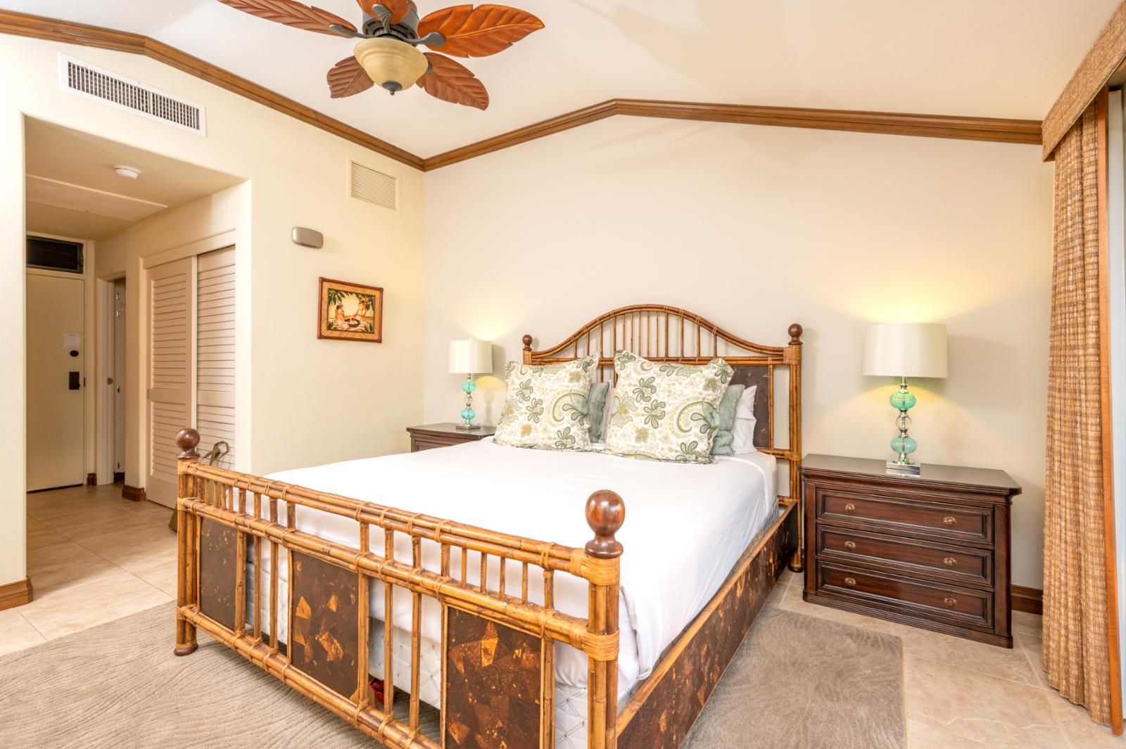 A bedroom with a wooden bed, two bedside tables with lamps, a ceiling fan, and a door leading to another room.