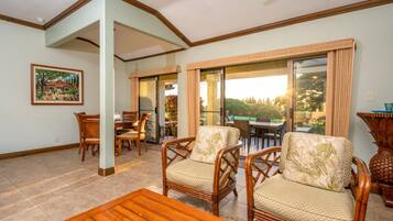 A spacious living area with a dining table, two wicker chairs, a wooden coffee table, and a large sliding glass door leading to an outdoor patio.