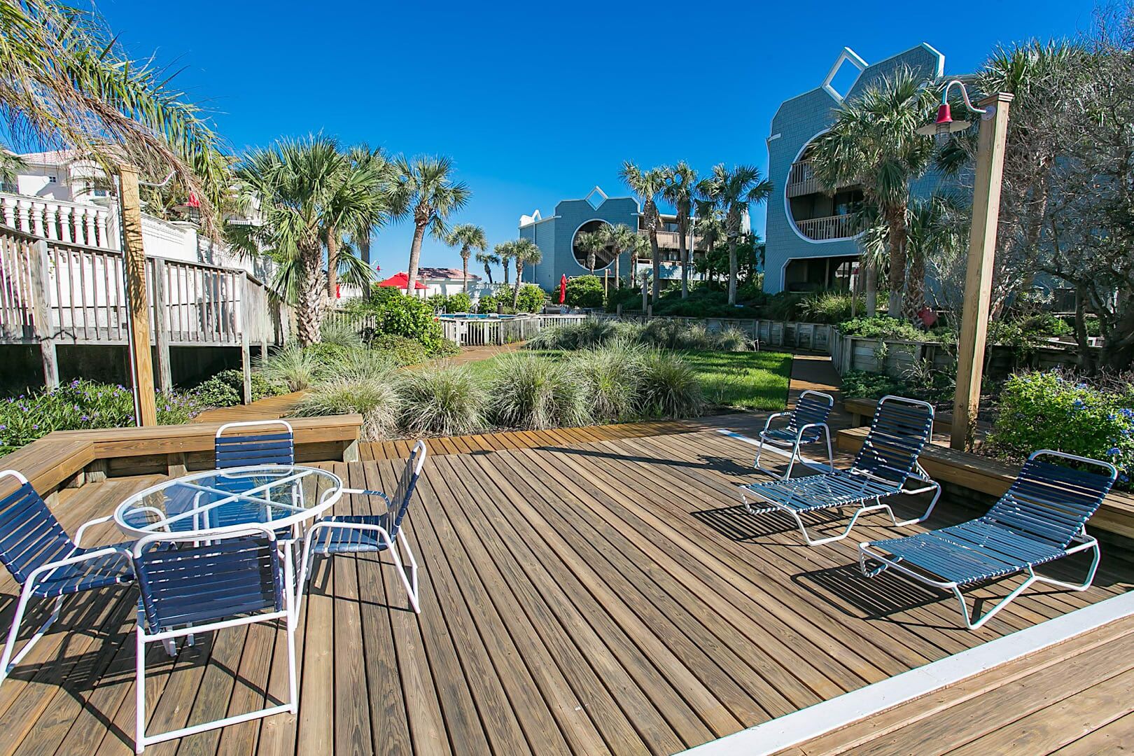 Une terrasse en bois avec des chaises longues et une table en verre, entourée de verdure et d’une piscine.