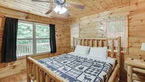 Quarto de cabana de madeira com cama, ventilador de teto e janela com cortinas.