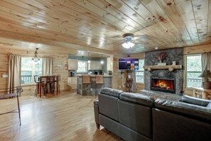 Un intérieur de chalet chaleureux avec une cheminée en pierre, un plancher en bois, un canapé, un coin repas et une cuisine.