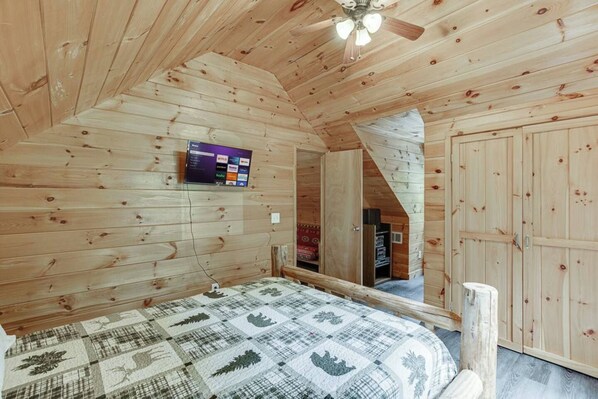 A wooden cabin bedroom with a bed, a ceiling fan, a mounted TV, and a wardrobe.